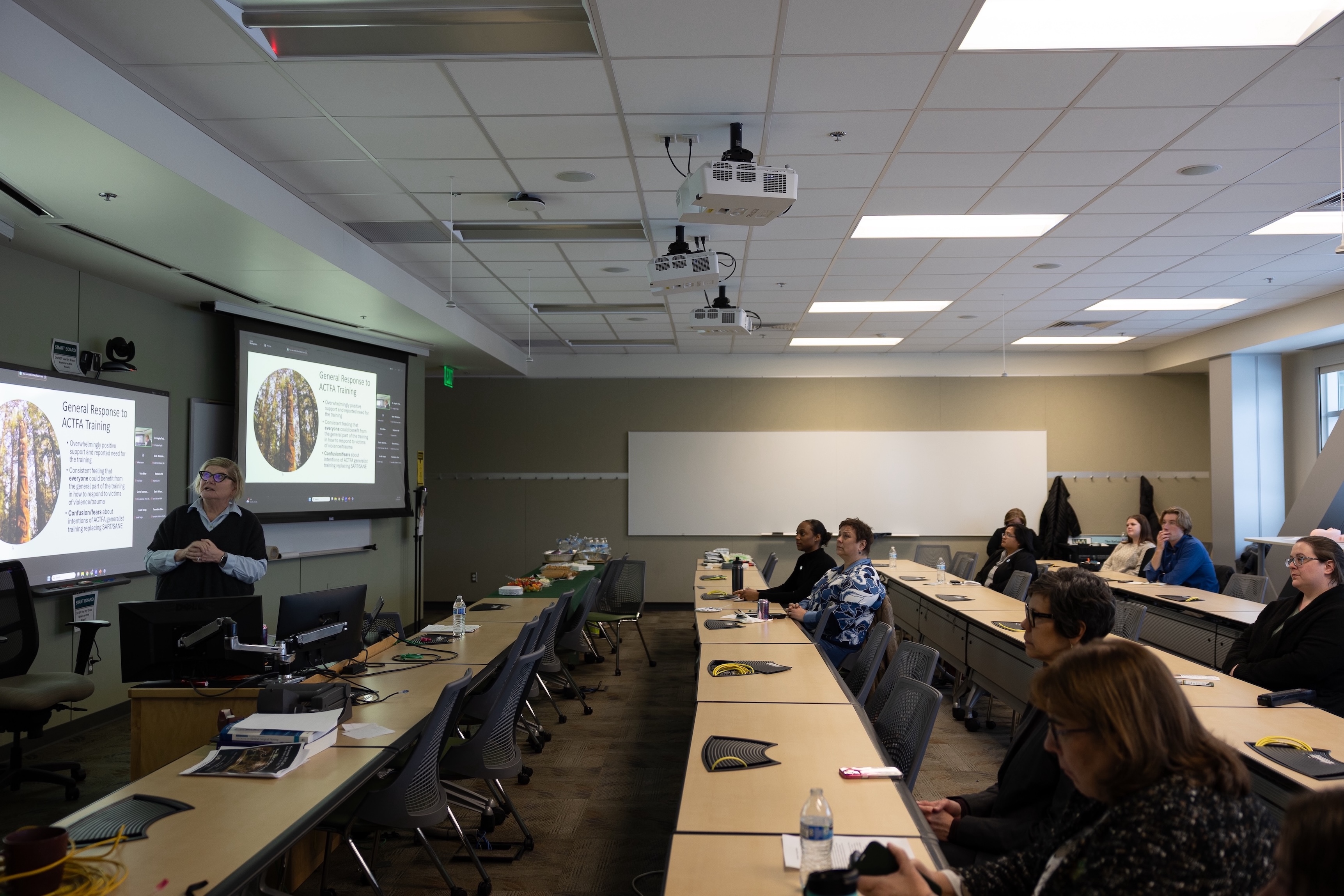Dr. Kathi Trawver stands in front of a projector screen in a classroom and presents. The classroom features rows of desk with audience members dispersted throughout.