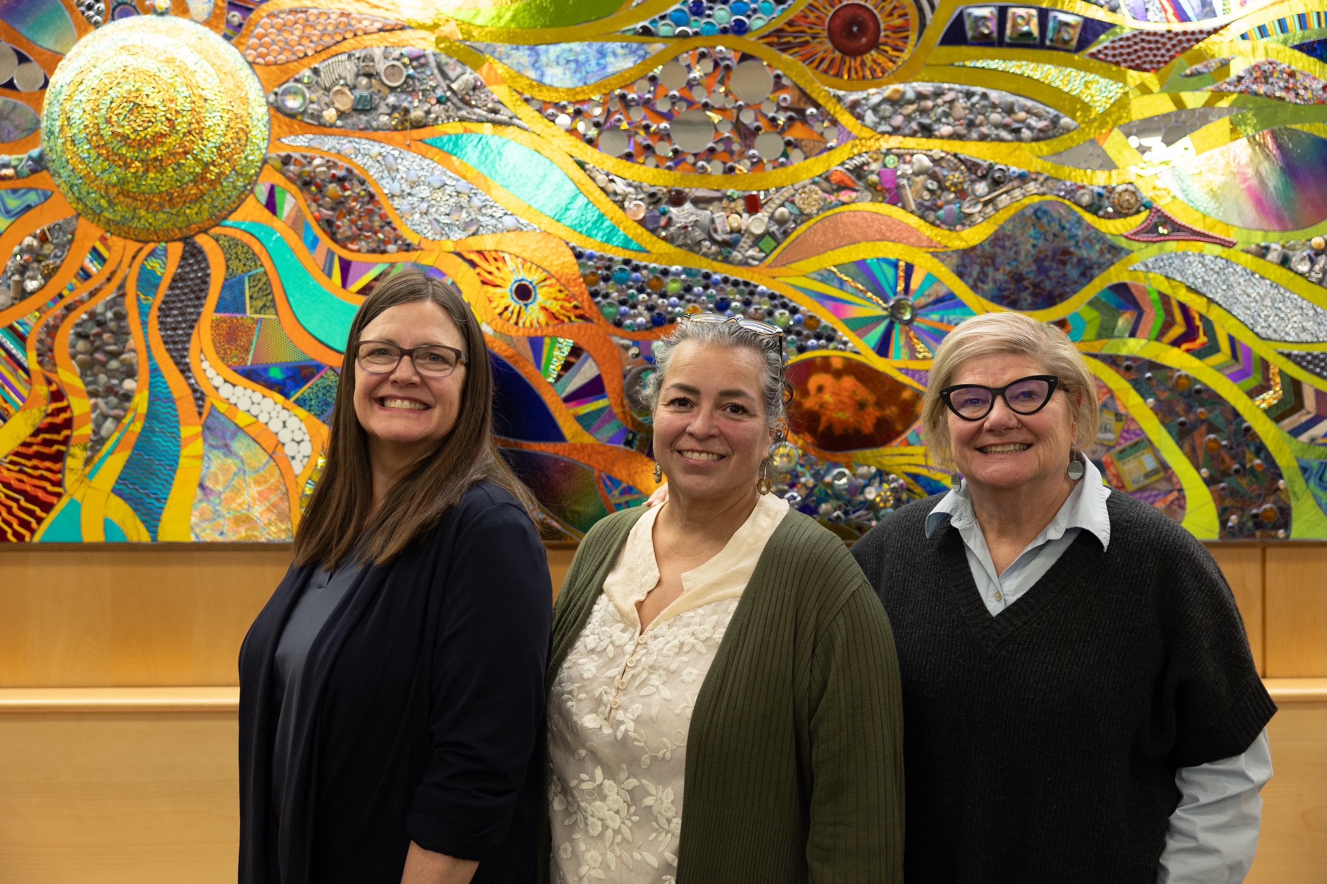 ACFTA team members Joanne Wiita, Angelia Trujillo, and Kathi Trawver stand in front of a mosaic, smiling.