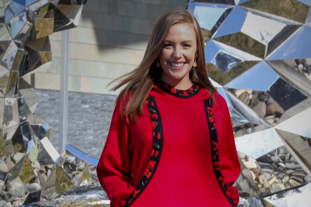 Laura Aspelund stands in front of the mirrored Heath Satow sculpture on the UAA campus. She is smiling and wearing a red kuspuk. 