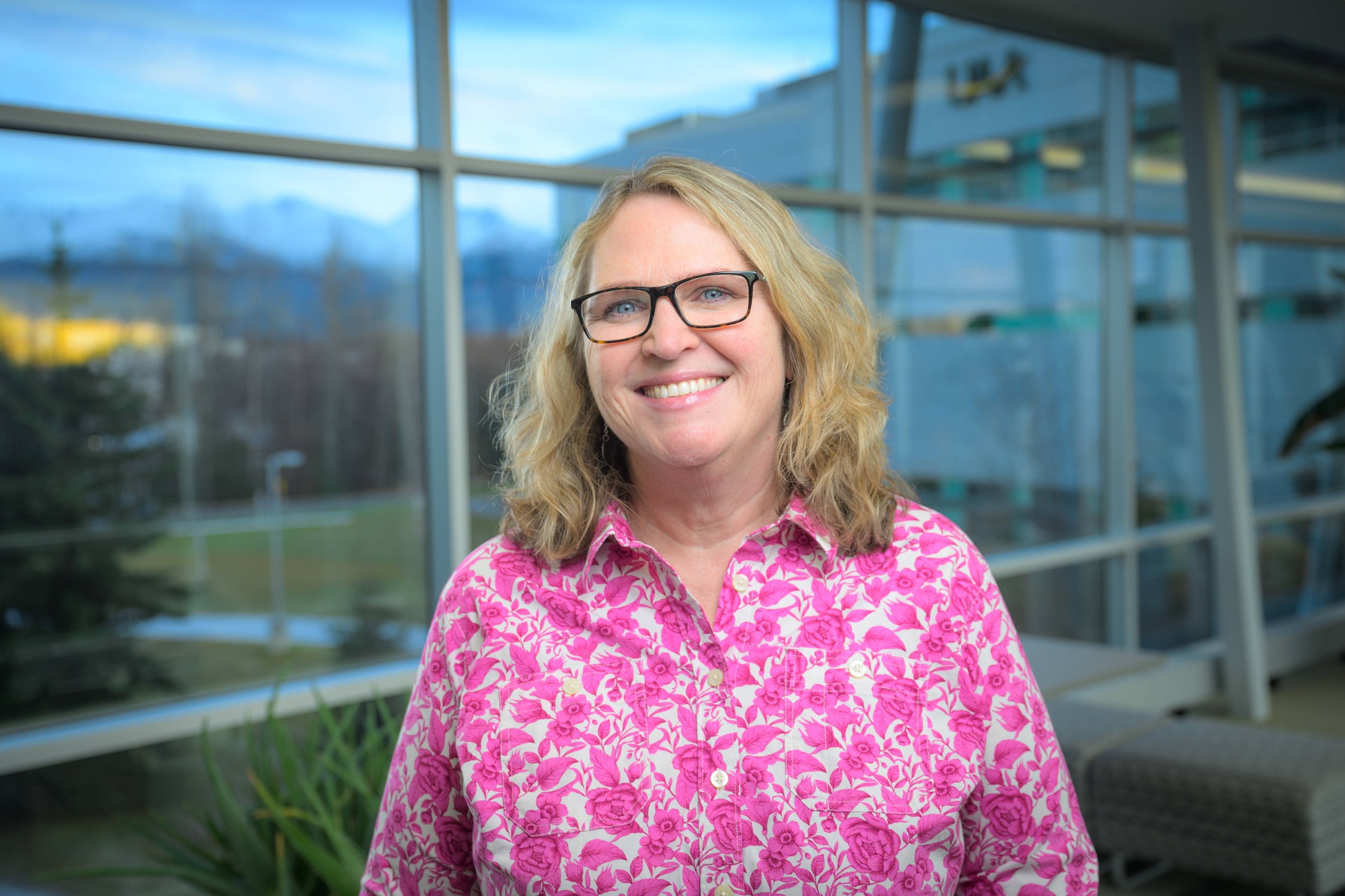 Stacy Brunquist stands in front of a wall of windows in the UAA Spine. She is wearing a pink and white patterned shirt and smiling at the camera.