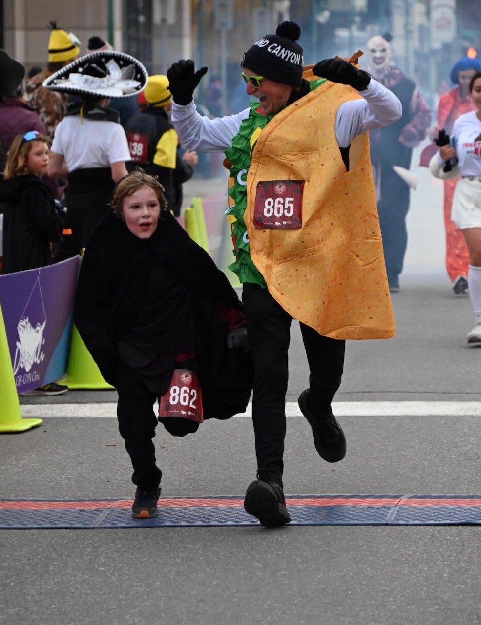 Jay Butler crosses the finish line of a race with his grandson. Butler is wearing a taco costime with a marathon bib pinned to it, and his arms are raised in celebration; his grandson is wearing a black costume.