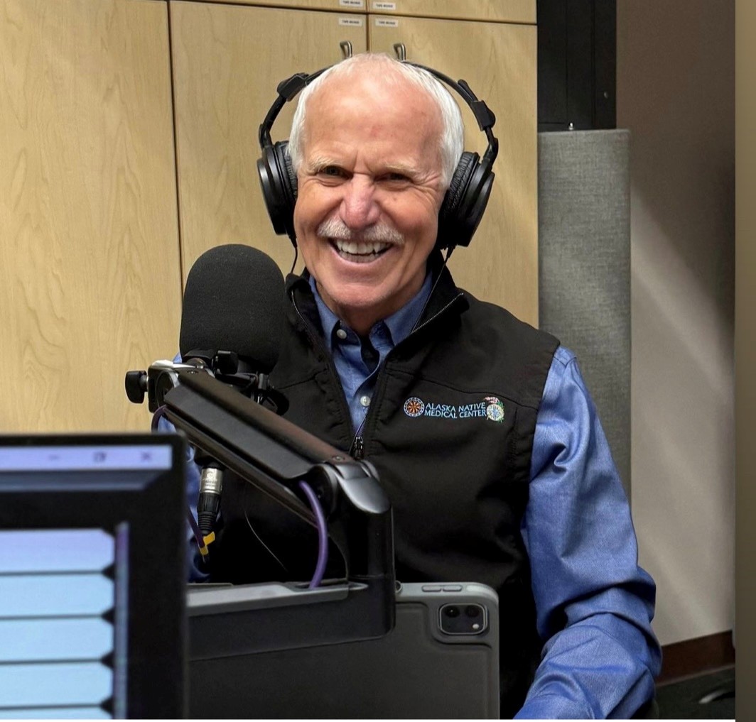 Dr. Jay Butler wears headphones and sits in front of a large microphone in a recording studio. He is smiling and wearing a blue button-down shirt under a black vest.