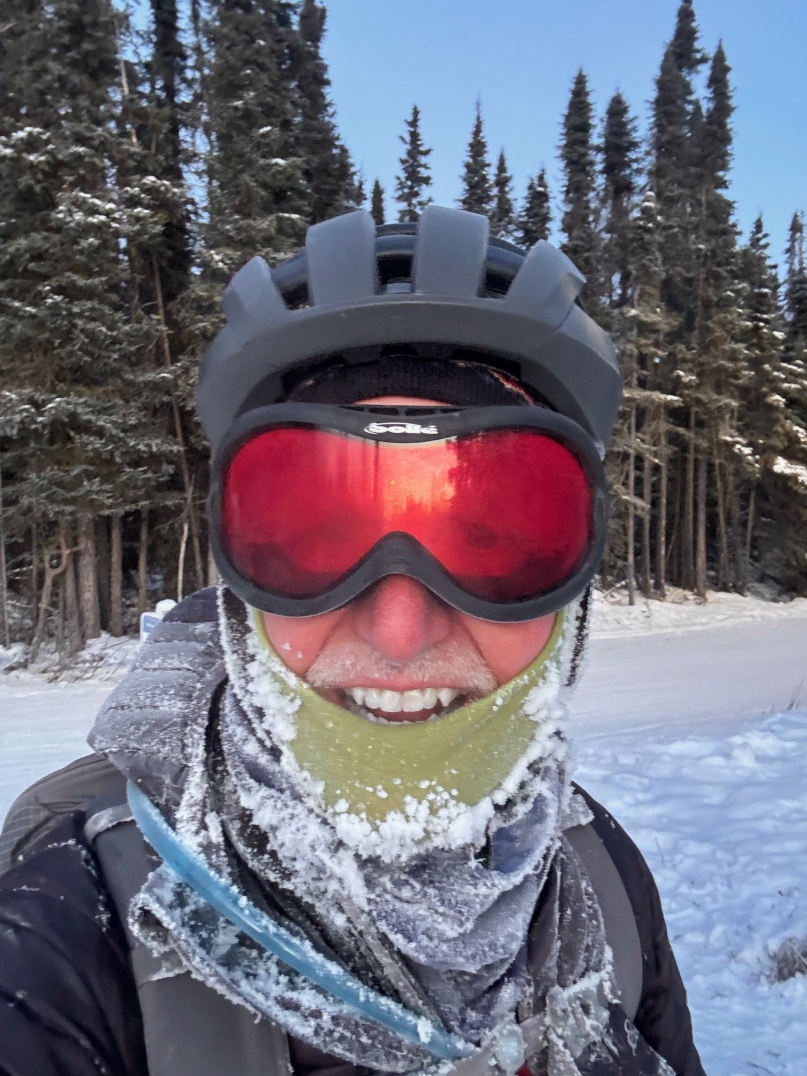 Jay Butler wears a bike helmet, goggles, and cold-weather outdoor gear. His neck gaiter and jacket are crusted with ice. Behind him is a snowy bike path and forest.