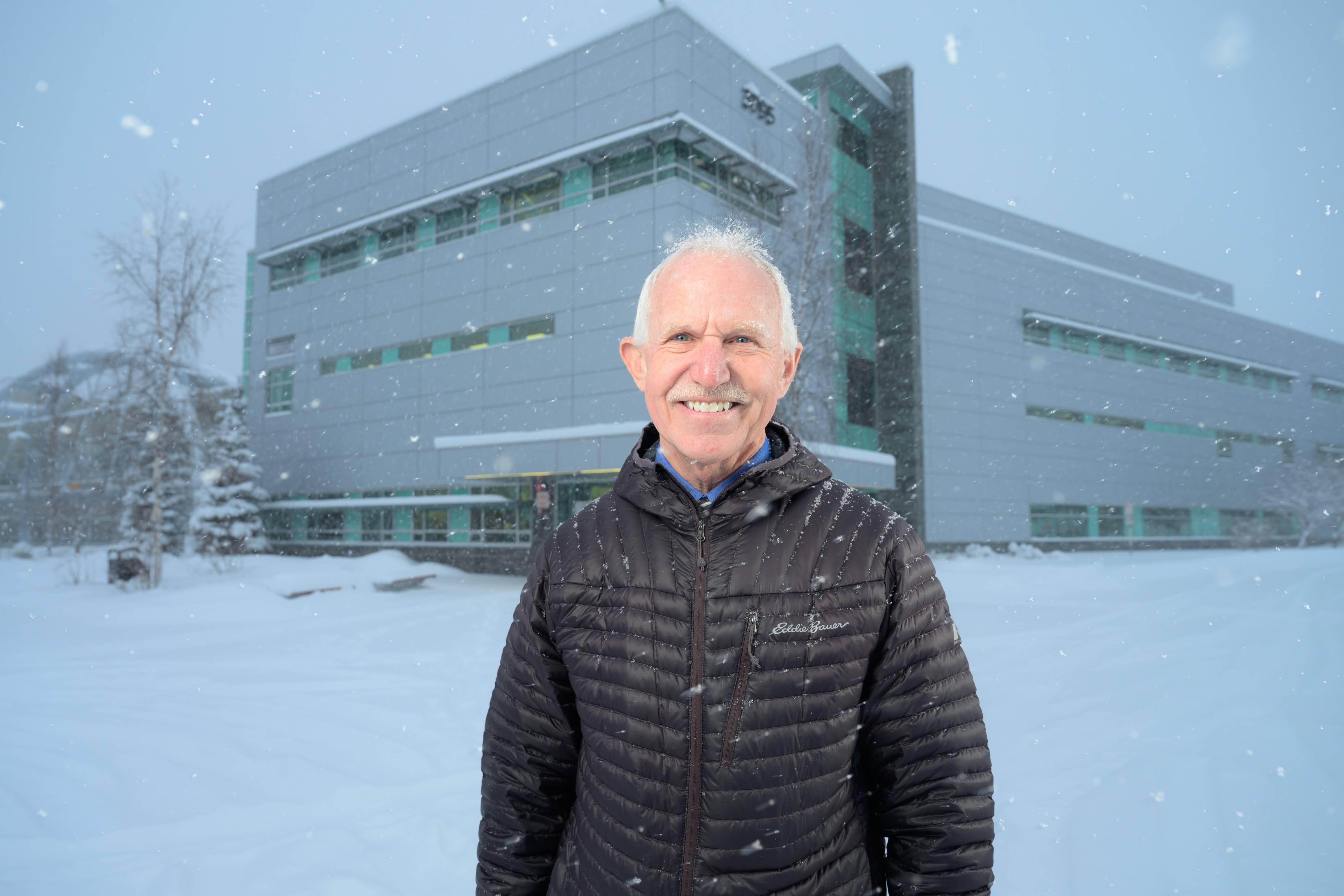 Jay Butler stands in front of a building at the UAA campus while it snows lightly. He is wearing a black puffy jacket and smiling. 