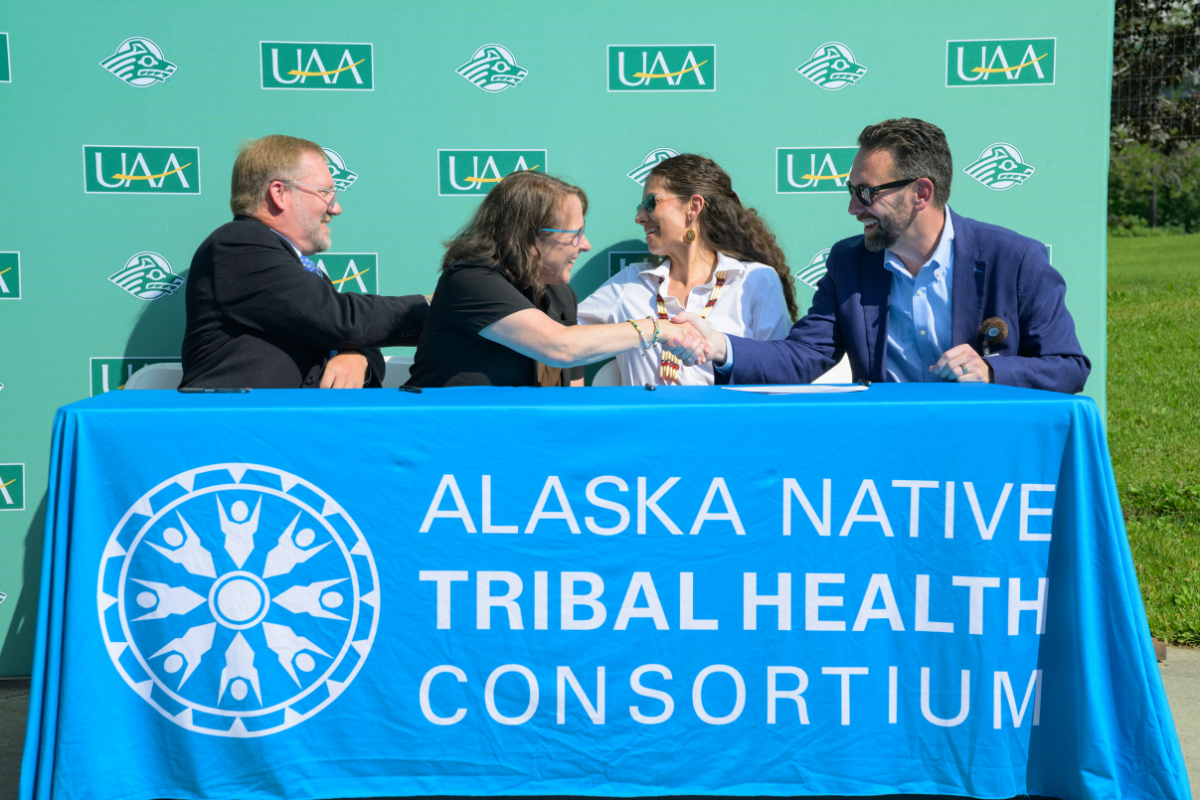 Four people seated on the same side of a rectangular table all shaking hands with one another