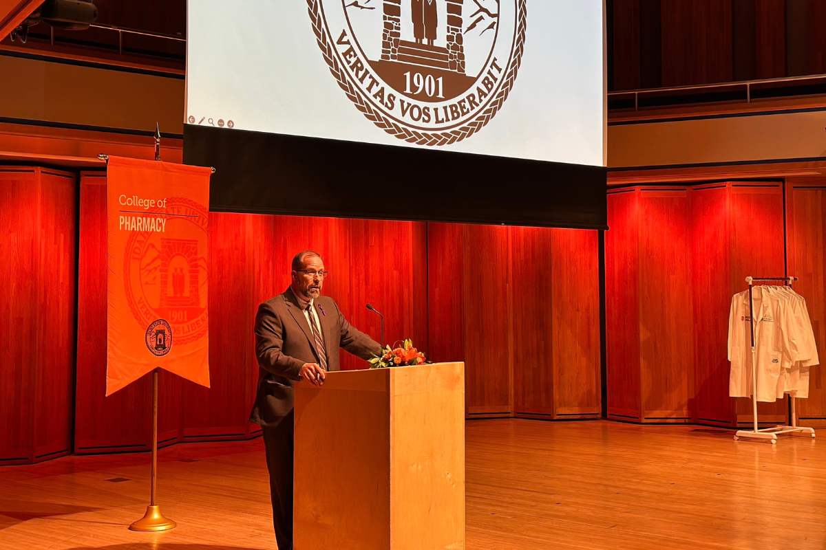 A man in a gray suit stands on stage behind a podium surrounded by orange lights, a rack of white coats, a projector with a school seal, and an orange banner that reads "College of Pharmacy"