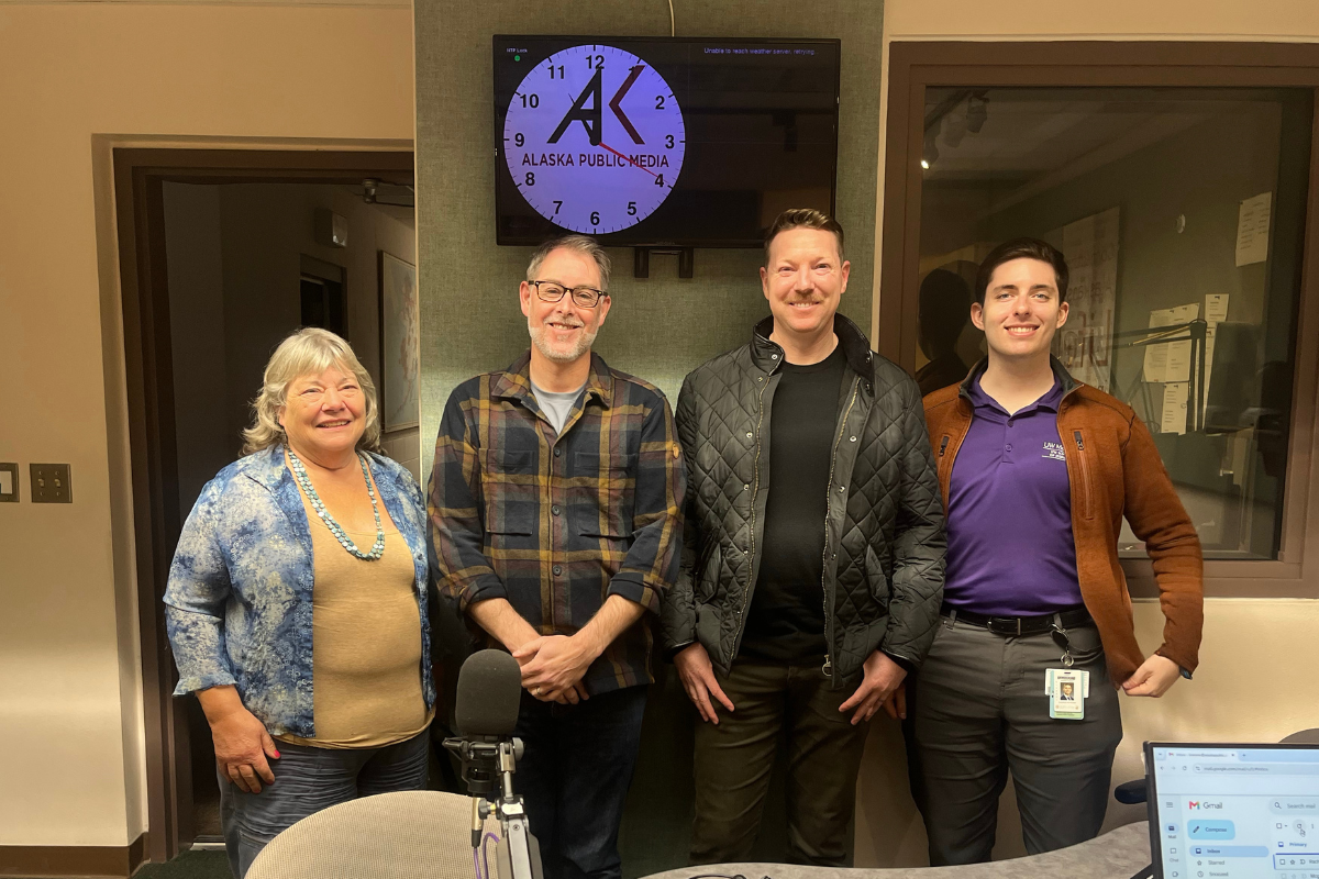 A woman and three men pose with a microphone in front of a clock