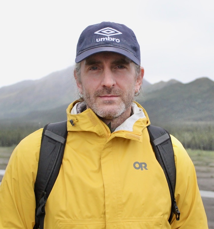 Shane in a yellow jacket standing in front of mountains