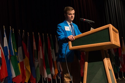 Student stands at a pondium at the head of a mock-UN assembly hall.