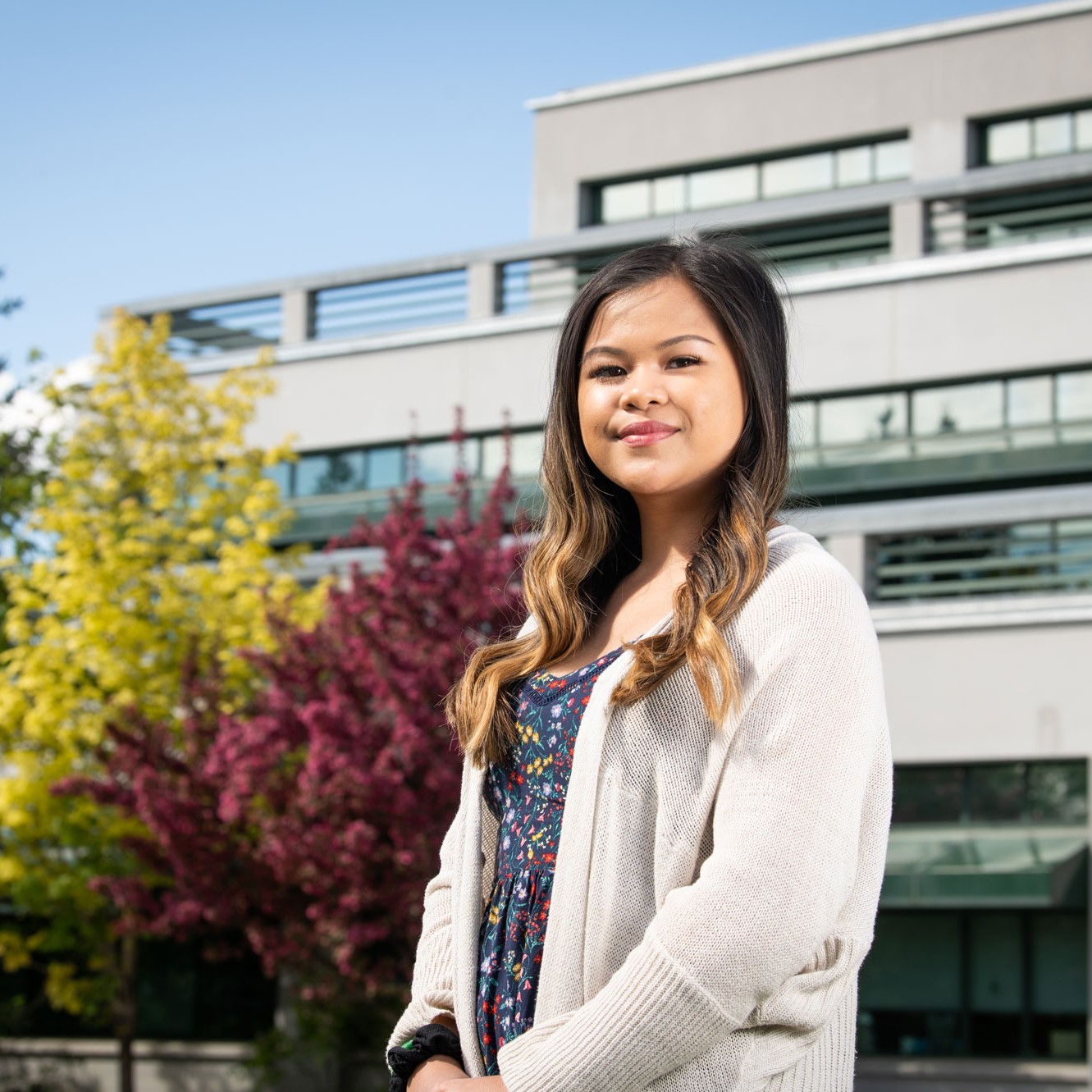 A woman in a white lab coat stands in front of a building and trees.