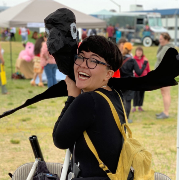 Woman in black clothing caries a papier-mache bird while on a grassy field.