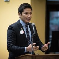 A man in a dark suit stands and speaks at a podium with a microphone.