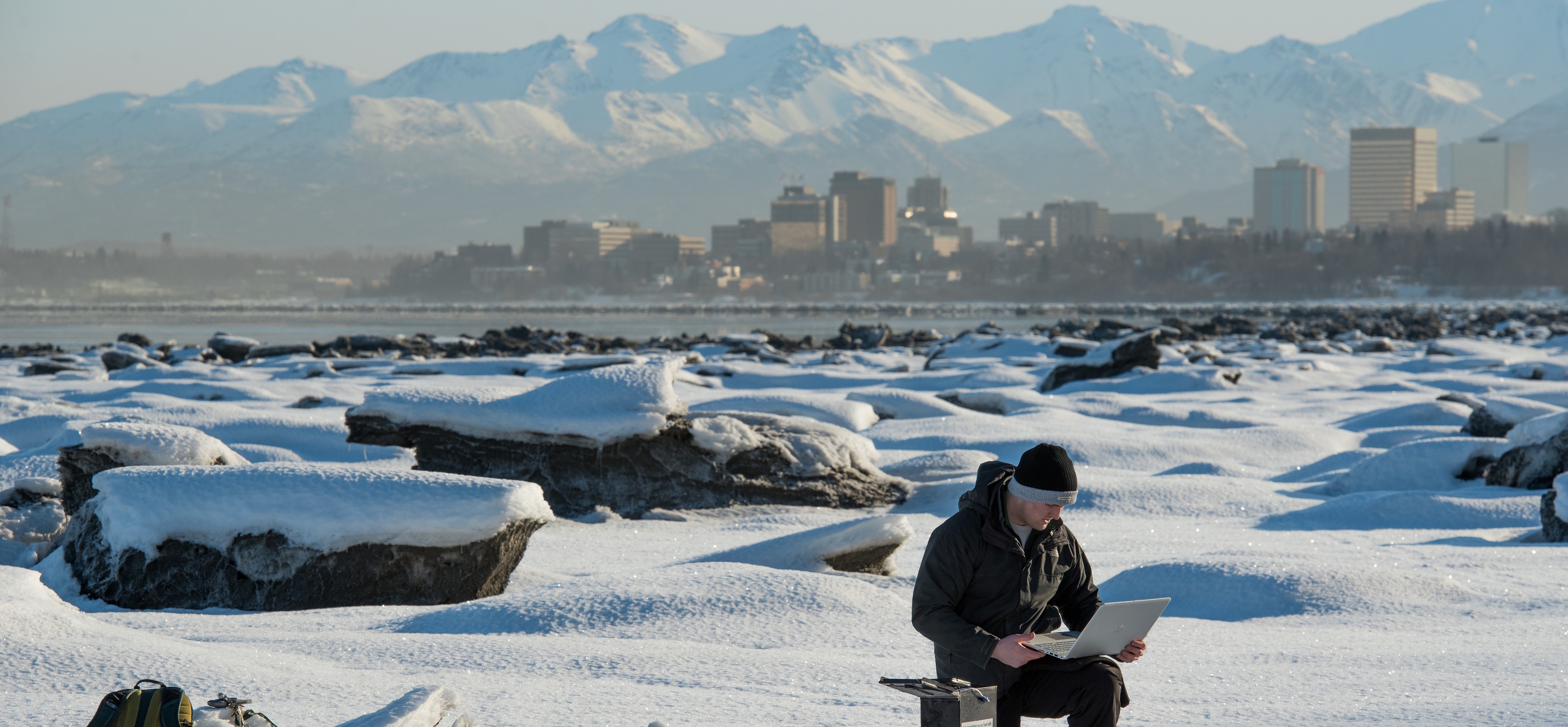 Student conducts research in the snow with Anchorage skyline and mountains in the background