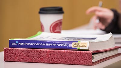 A behavior analysis textbook sits on a binder next to a coffee cup