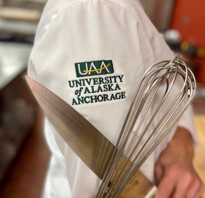 A knife and whisk held against University of Alaska Anchorage embroidery on a chef jacket