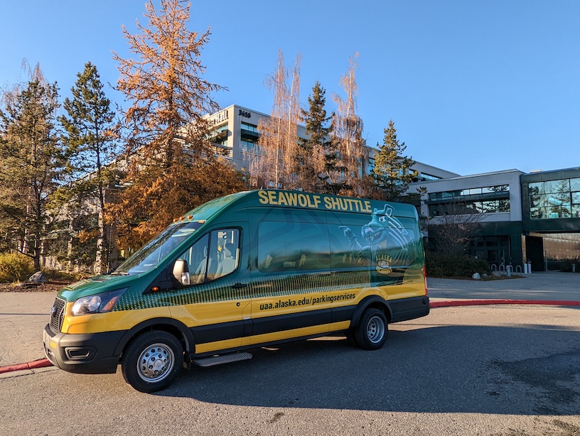 The Seawolf Shuttle parked against the sidewalk, outside UAA. There are blue skies, and it appears to be in the fall. 