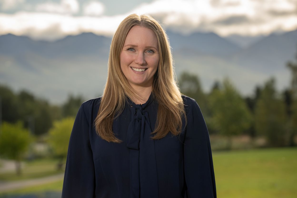 A woman with long blonde hair is smiling outdoors with a scenic mountain landscape behind her.