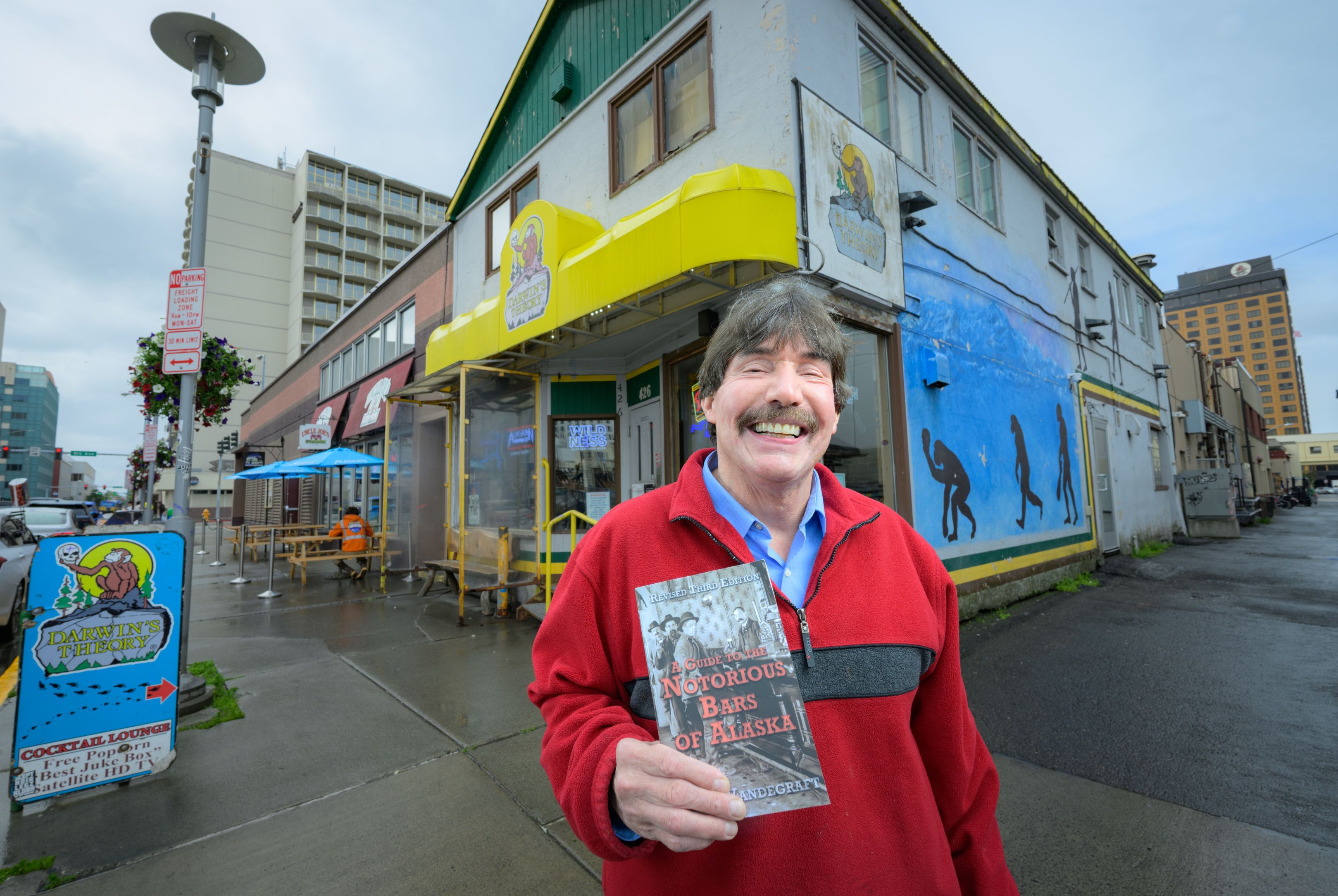 A man in a red jacket stands in front of a bar, holding a book titled "A Guide to the Notorious Bars of Alaska".