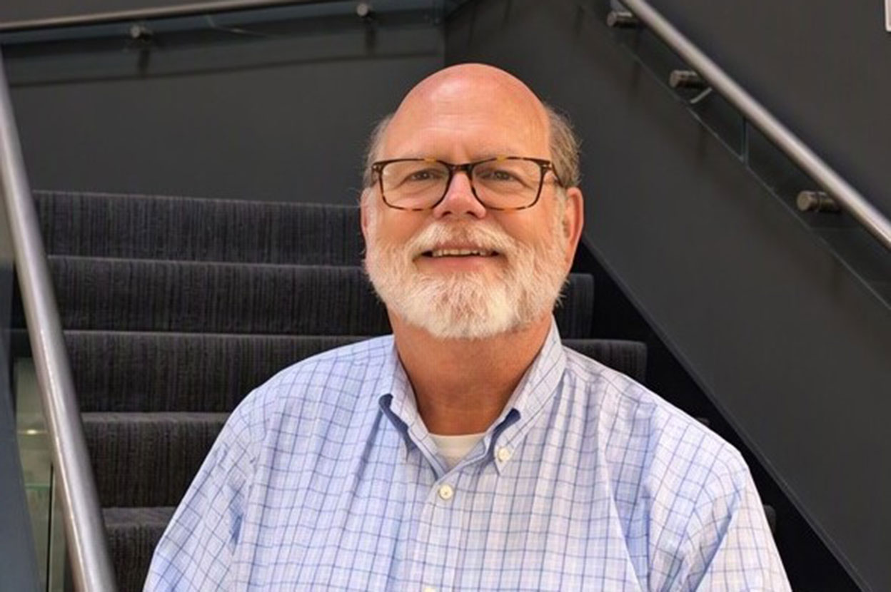 A man wearing glasses and a button-up shirt stands smiling at the bottom of a stairwell.
