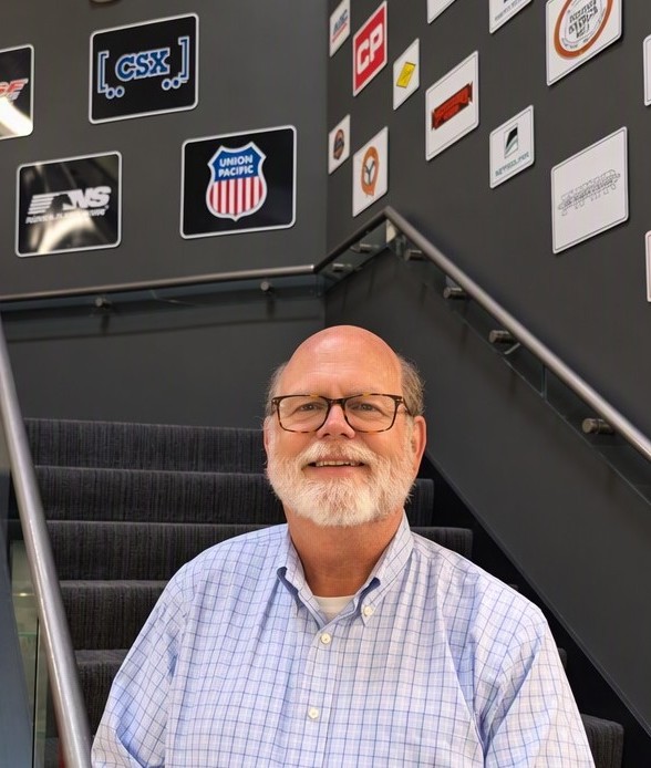 A man wearing glasses and a button-up shirt stands smiling at the bottom of a set of stairs.