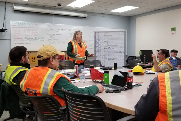 Management and facilities teams met in a classroom to discuss plans for earthquake recovery. We see one team member standing in front of the whiteboard, speaking. Everyone is wearing orange safety vests.