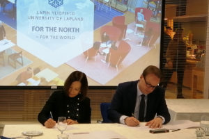 UAA Chancellor Cathy Sandeen sits at a table with Antti Syväjärvi, rector of the University of Lapland in Finland. They are signing a memorandum of understanding. Behind them is a projector screen with the University of Lapland logo.