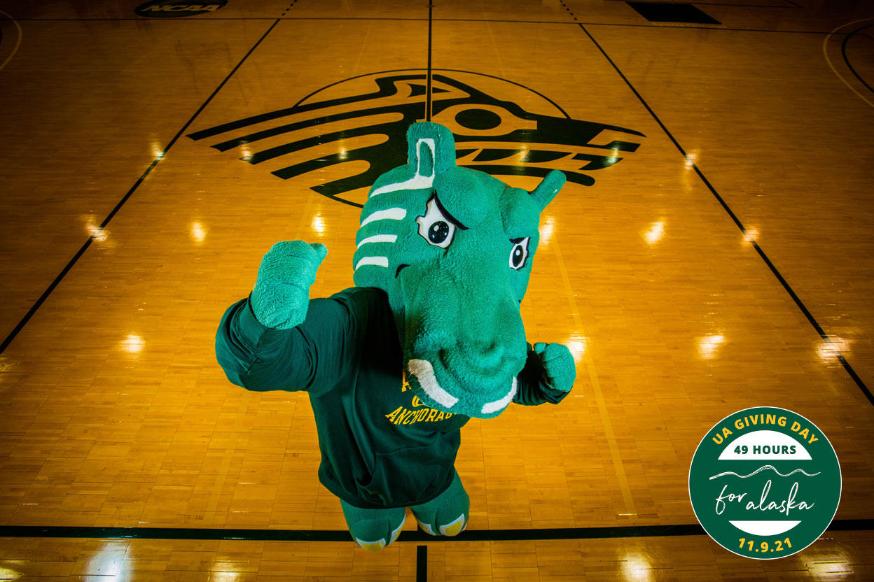 Spirit jumps up in celebration on the court at the Alaska Airlines Center. Giving Day logo is in lower-right corner. (Photo by James Evans / UAA)