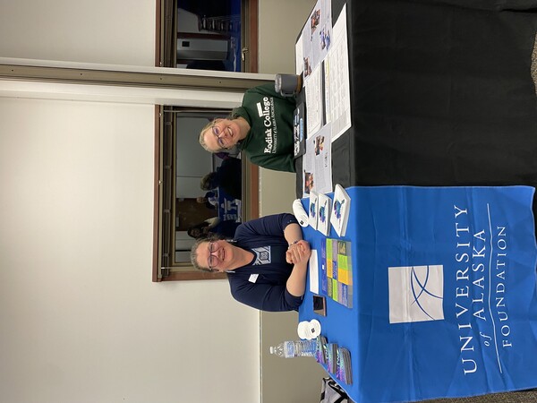 two women sit behind a booth, with blue table two women sit behind a booth, with blue table cover that reads "UA Foundation"