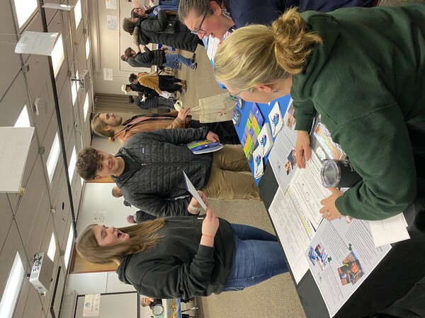 a male and female student stand in front of a table, with two women behind the table talking to them