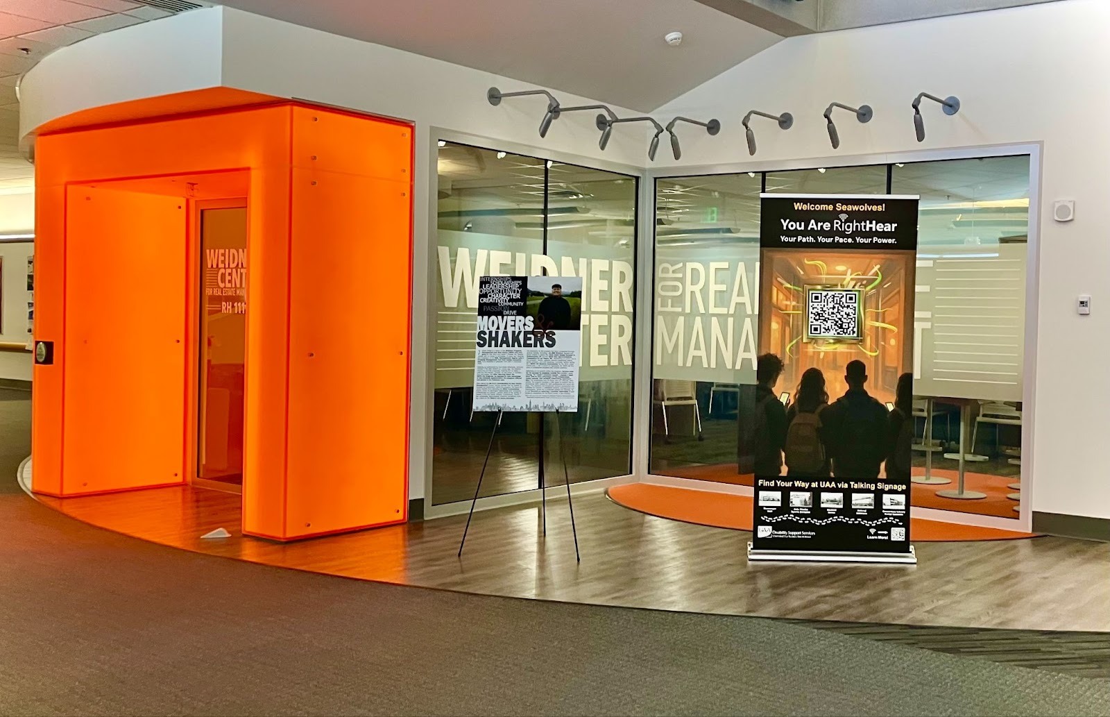 RightHear educational banner with a glowing QR code displayed in the UAA Rasmuson Hall lobby near the Weidner Center, featuring a diverse group scanning the code with smartphones.