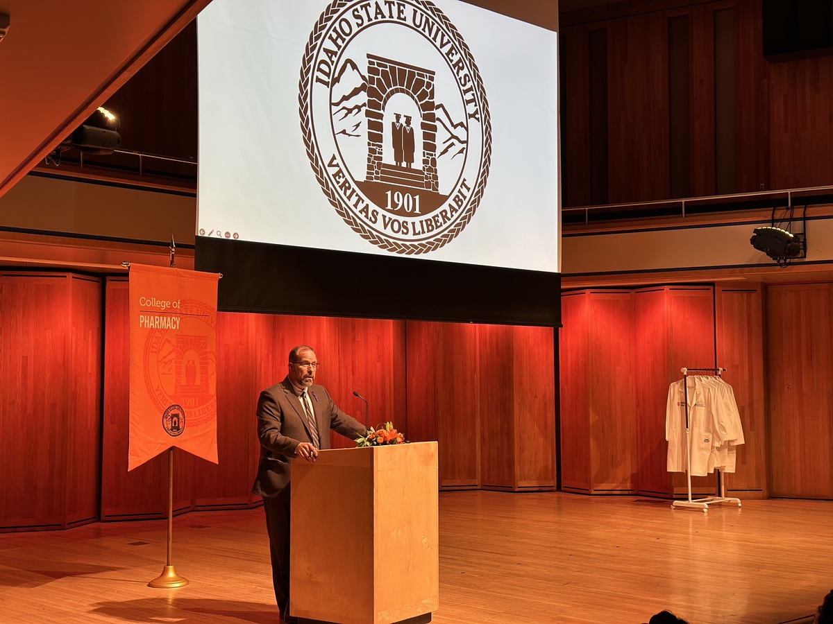 Man speaking at podium with Idaho State University banner behind him and rack of white coats to his left