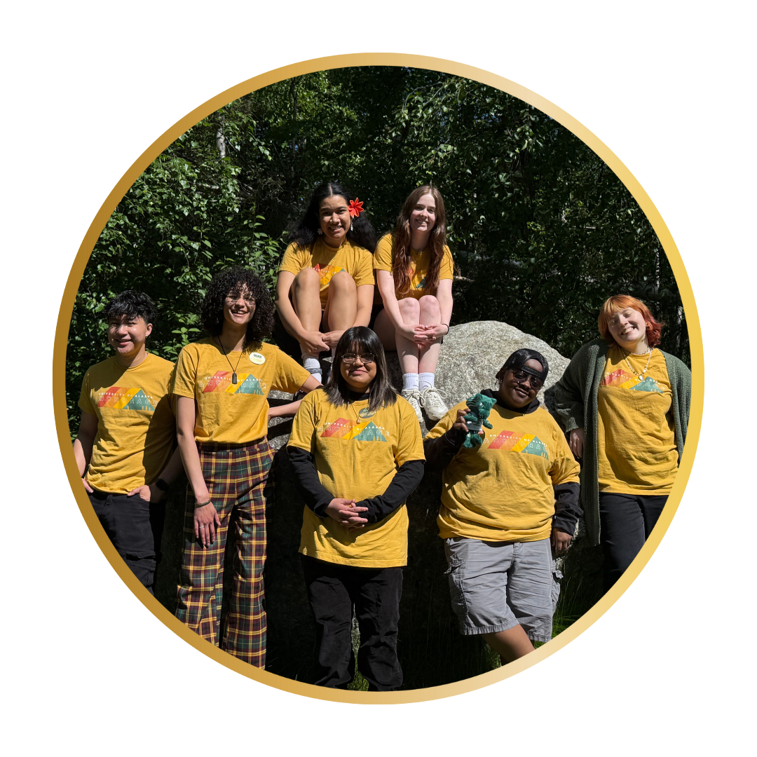 Group portrait with six student orientation leaders during the summer in Alaska. It is a beautiful sunny day. The background is surrounded by green trees and grass. They are also sitting and leaning on a big rock
